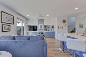 Una sala de estar moderna con un sofá gris, una cocina con armarios blancos y una zona de bar con taburetes blancos.