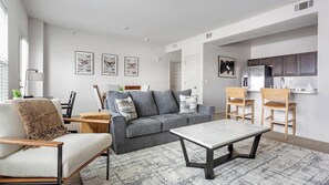 Una sala de estar moderna con un sofá gris, una silla de madera, una mesa de centro de mármol y una zona de cocina con armarios y un refrigerador.