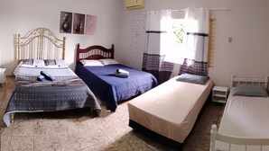 A room with three beds, a window with curtains, a wall-mounted air conditioner, and a small bedside table.