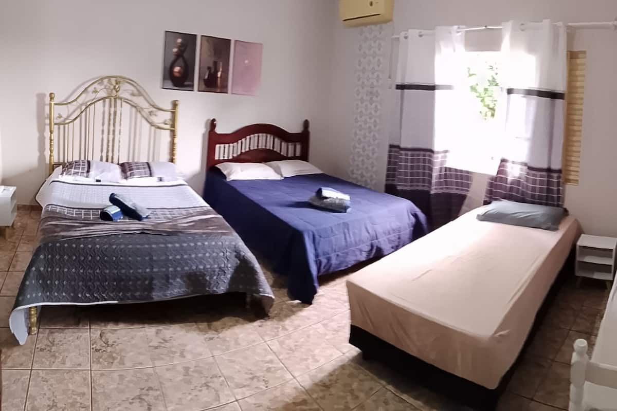 Una habitación con tres camas, una ventana con cortinas, aire acondicionado montado en la pared y una mesita de noche pequeña.