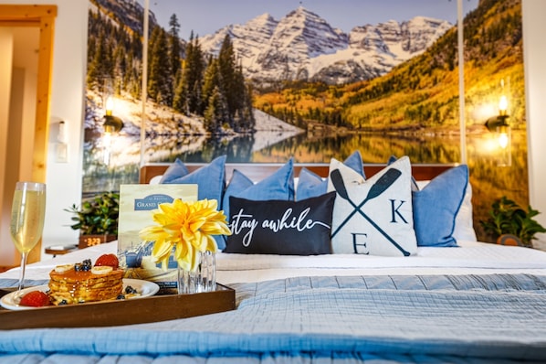 A bed with a mountain view mural, a tray with pancakes and berries, a glass of champagne, and a card.