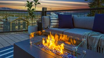 A balcony with a glass table holding a fire feature, a sofa with cushions, and a potted plant.
