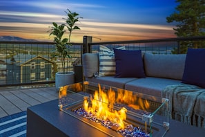 A balcony with a glass table holding a fire feature, a sofa with cushions, and a potted plant.