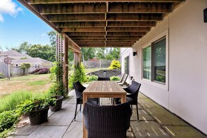 A covered patio with a wooden table and chairs, potted plants, and a view of the backyard.