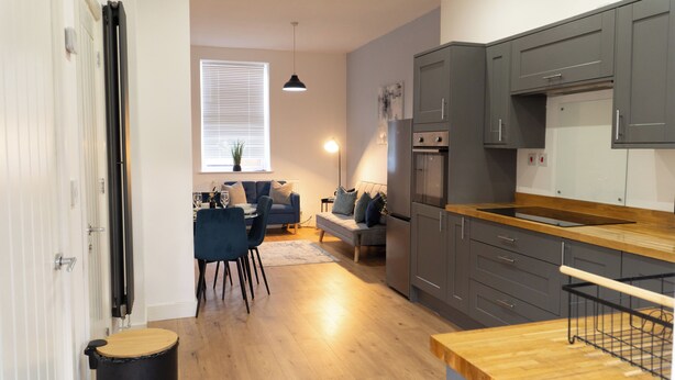 Une cuisine moderne avec des meubles gris, un plan de travail en bois et un coin repas comprenant des chaises bleues et un canapé gris.