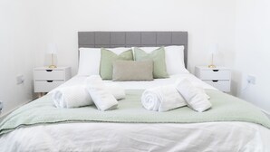 Un lit bien fait, avec une tête de lit grise, du linge de lit blanc et des coussins verts. On peut voir deux tables de chevet avec des lampes, et une prise de courant est visible sur le côté gauche du mur.