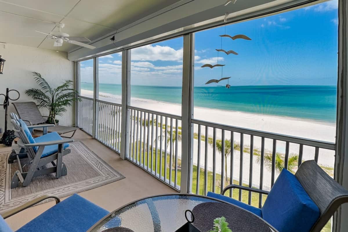 A balcony with a view of a beach, a chair, a table, and a hanging bird ornament.