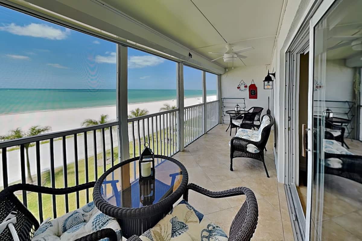 A balcony with a view of the beach, wicker furniture, a table, and a chair.