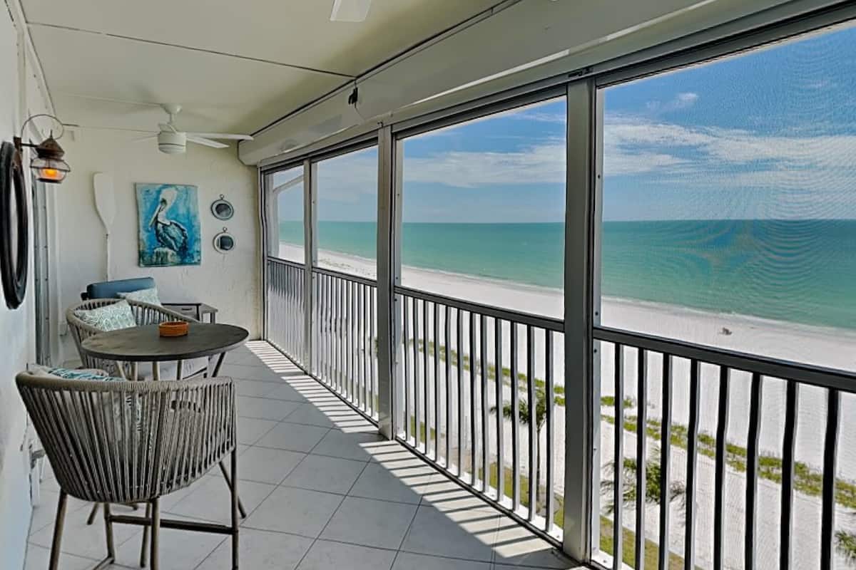 A balcony with a view of the beach, a table with chairs, and a painting on the wall.