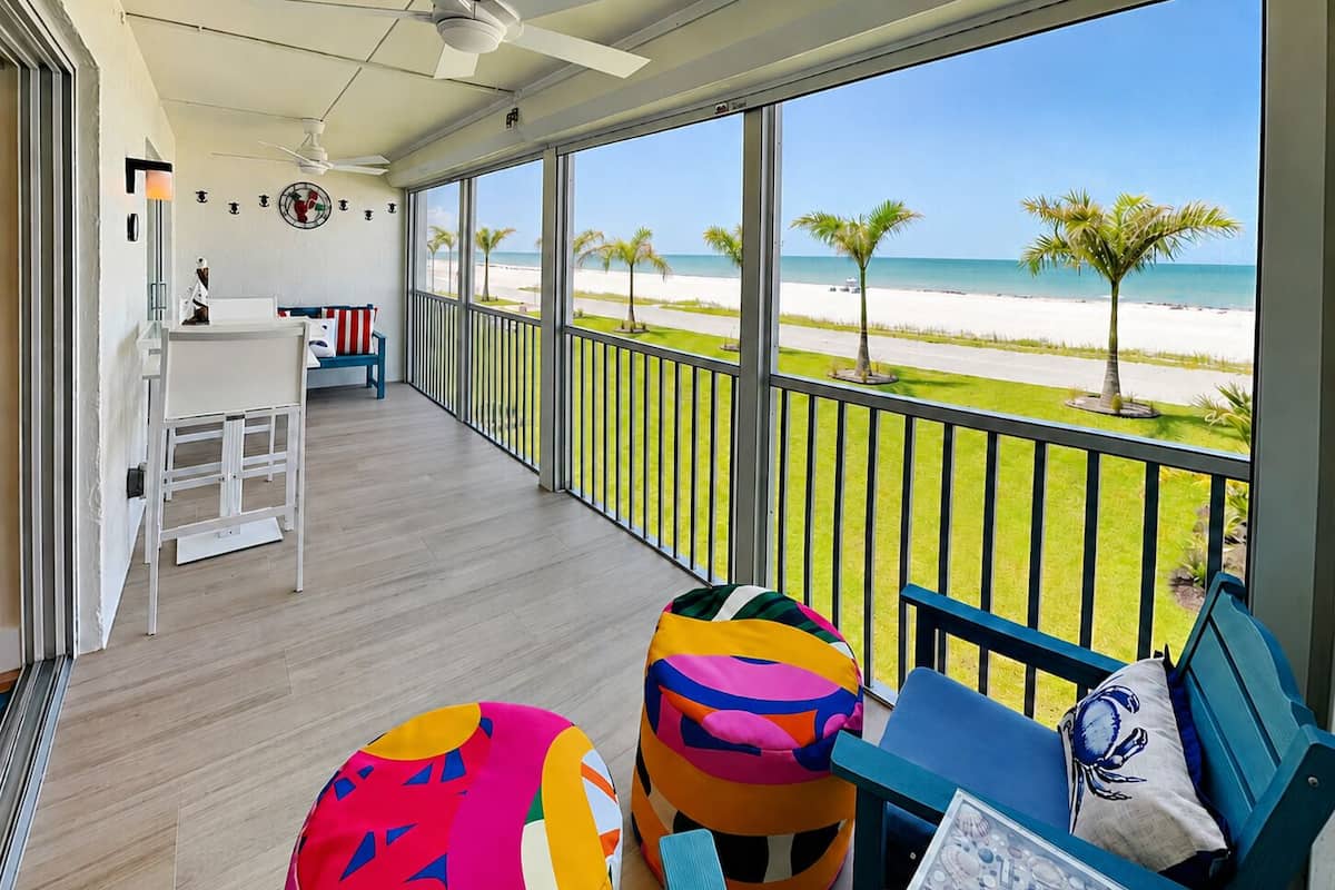 A balcony with a view of the beach, a white desk, colorful bean bags, and a blue sofa with a decorative pillow.