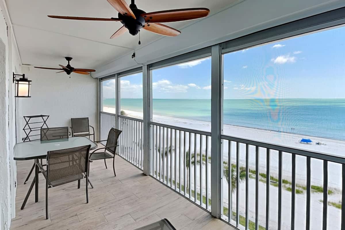 A balcony with a view of the beach, ceiling fans, and a table with chairs.