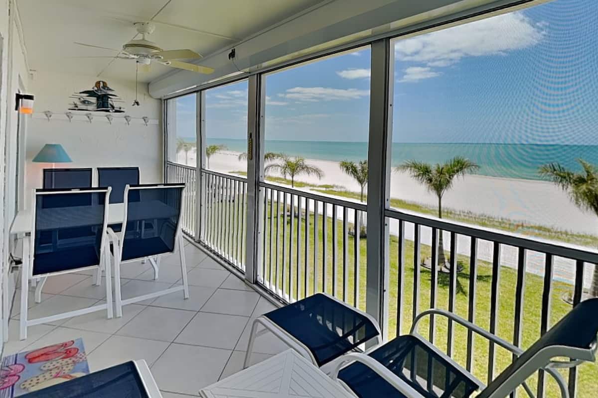 A balcony with a view of the beach, a ceiling fan, and a table with chairs.