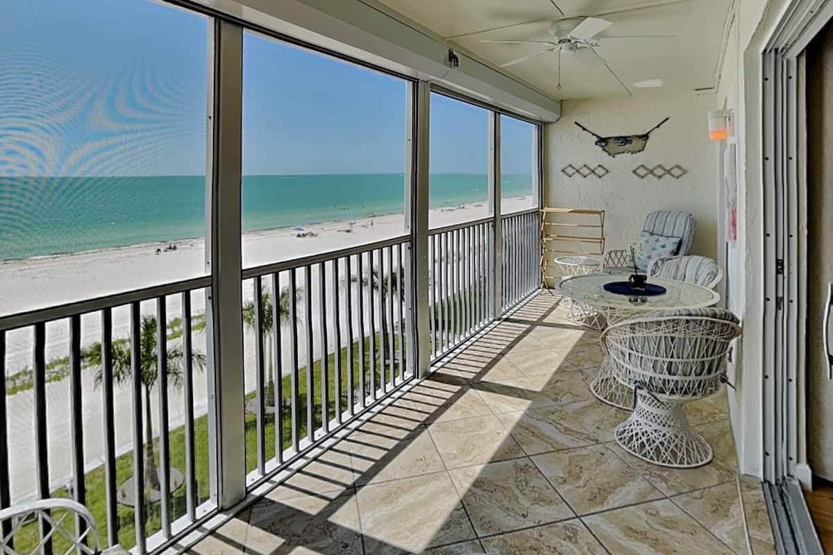 A balcony with a view of the beach, a ceiling fan, and a table with chairs.