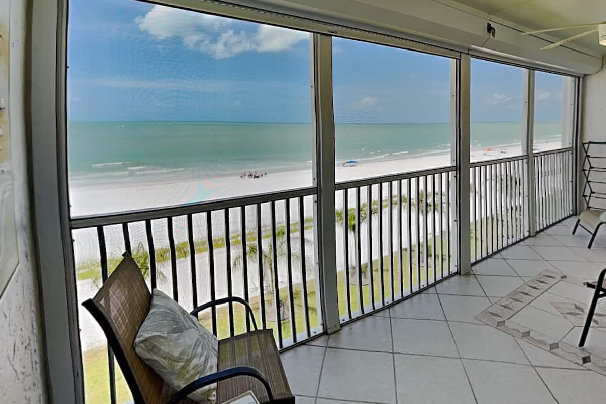 A balcony with a view of the beach, a chair, and a table.