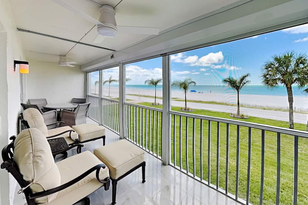 A balcony with a view of the beach, a table with chairs, and a ceiling fan.