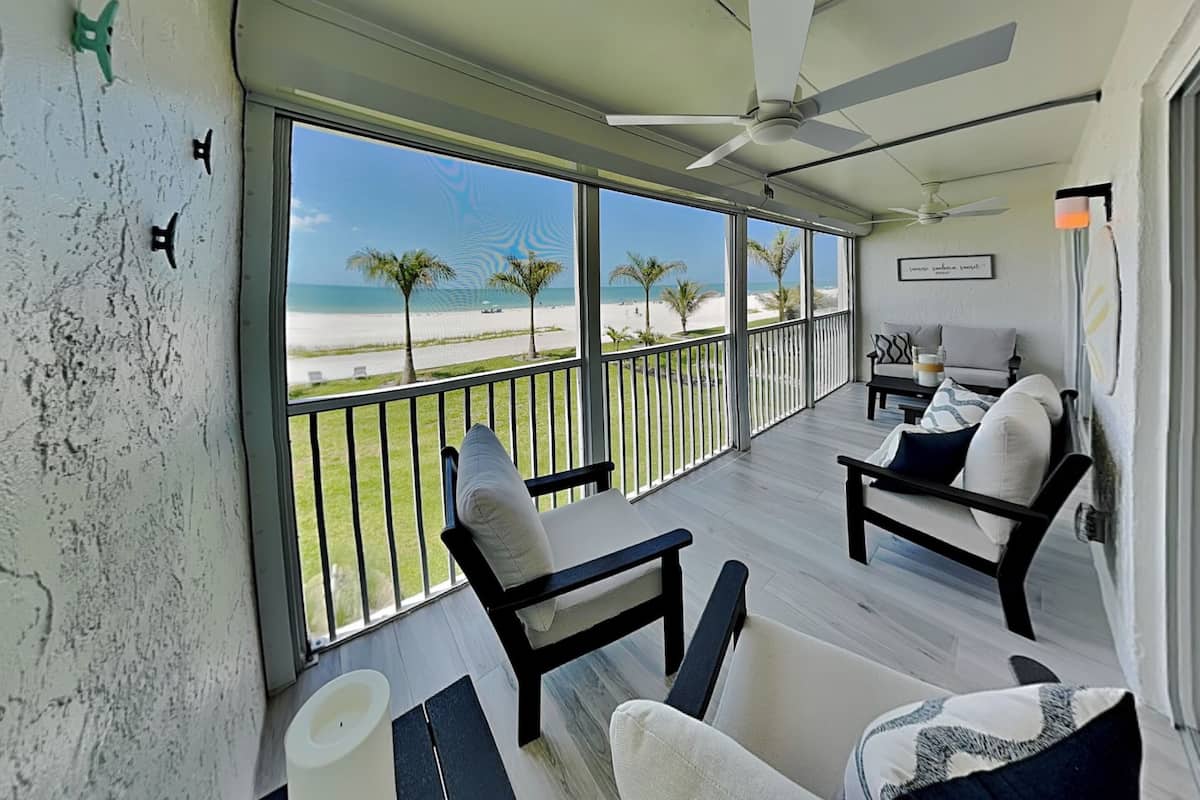 A balcony with a ceiling fan, a view of the beach, and a seating area with chairs and a table.