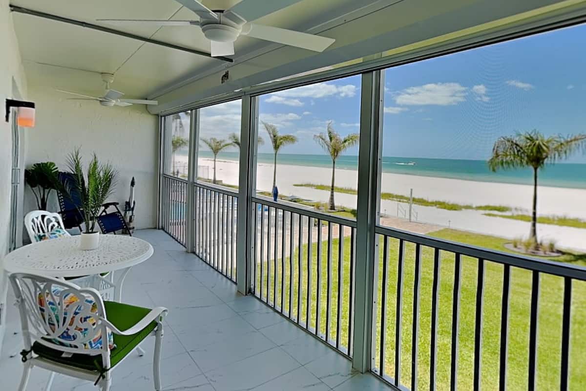A balcony with a view of the beach, a ceiling fan, and a table with chairs.