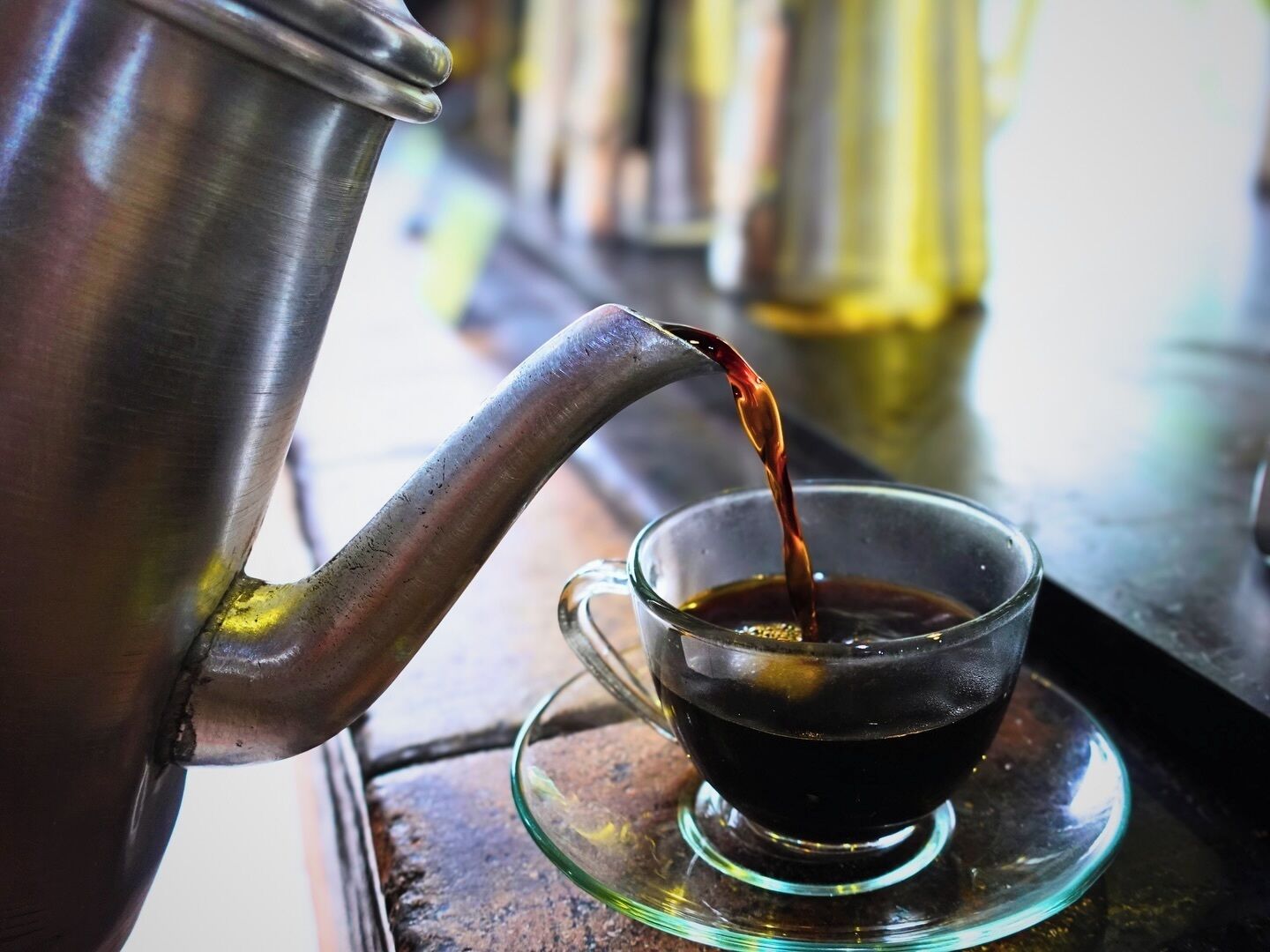 A metal coffee pot pouring liquid into a clear glass cup on a saucer.