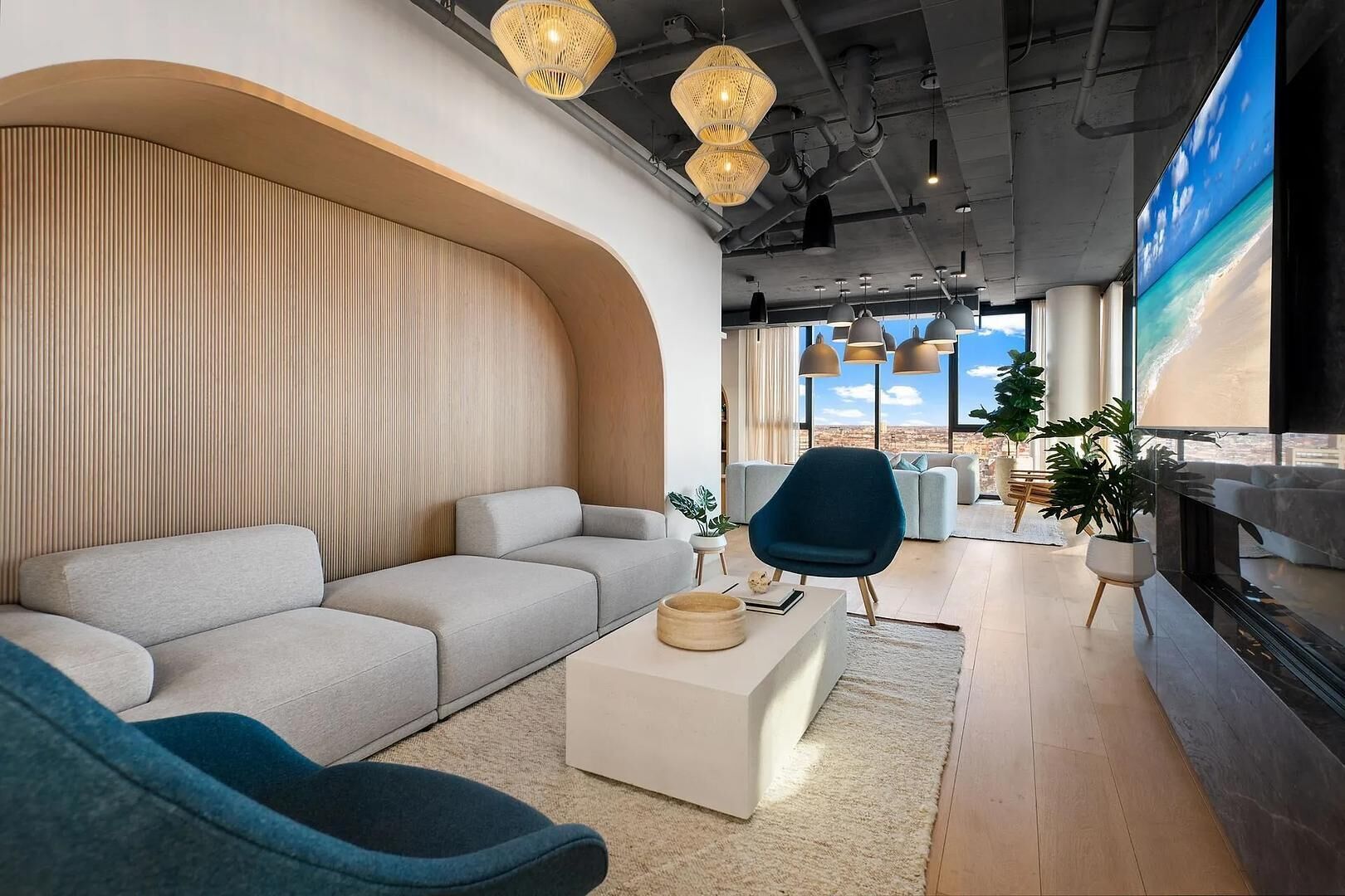 Een moderne woonkamer met een bank, een blauwe fauteuil, een salontafel en een grote tv die een strandscène toont.