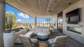 A rooftop terrace with a sectional sofa, round coffee table, wicker chairs, a large potted plant, and a flat-screen TV. - Landing | Beautiful 1BD, Yoga Studio, Pool (Chicago)