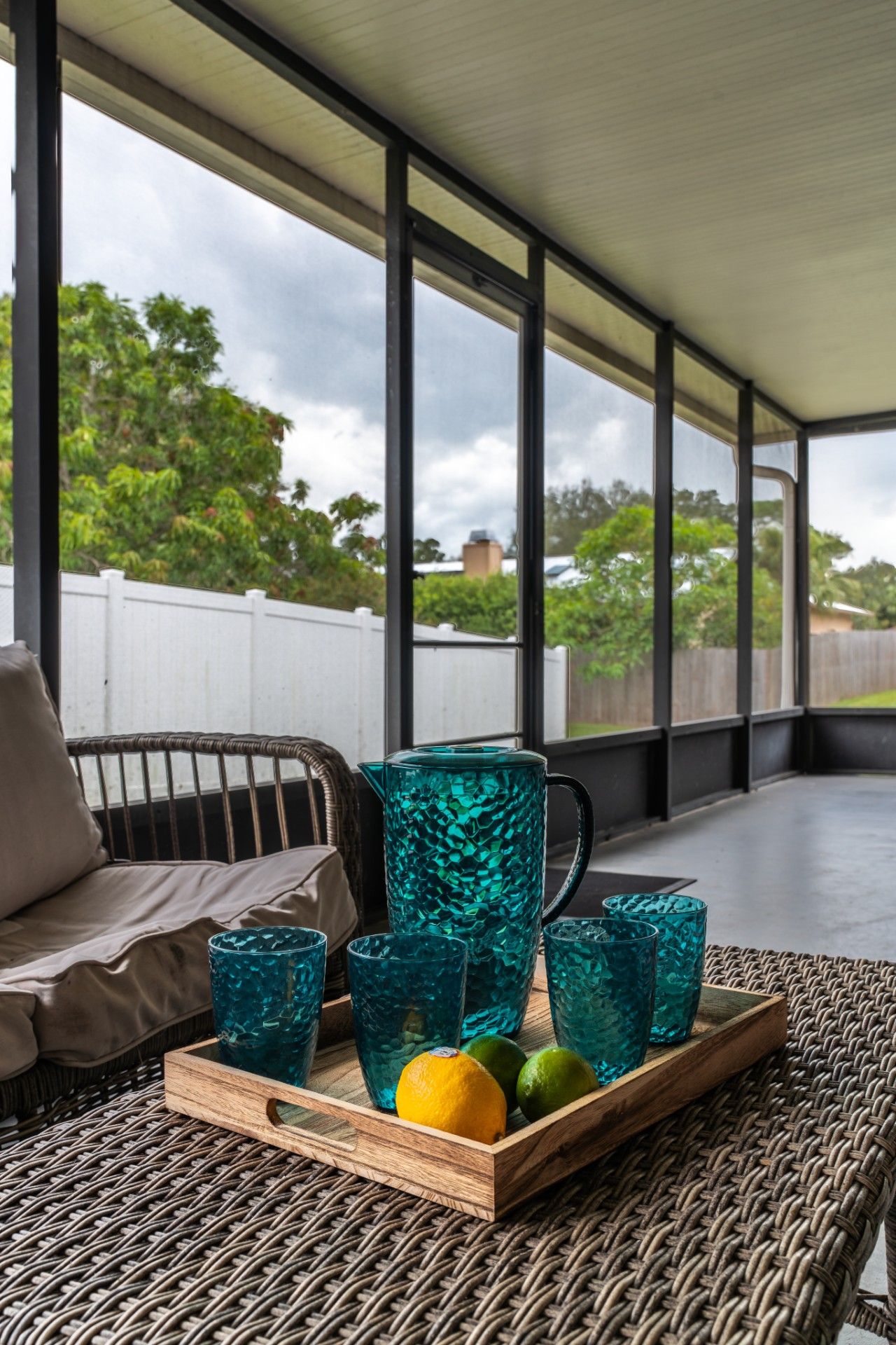 A covered outdoor seating area with wicker furniture, a wooden tray with blue glassware, and a view of the sky and trees.
