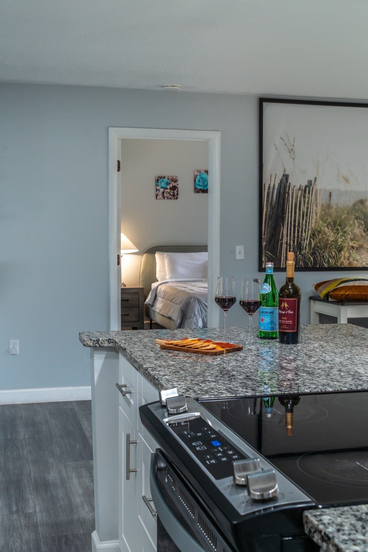 A kitchen with a built-in stove, a countertop with wine glasses and bottles, and a view into a bedroom with a bed and artwork.
