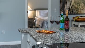 A kitchen with a built-in stove, a countertop with wine glasses and bottles, and a view into a bedroom with a bed and artwork.