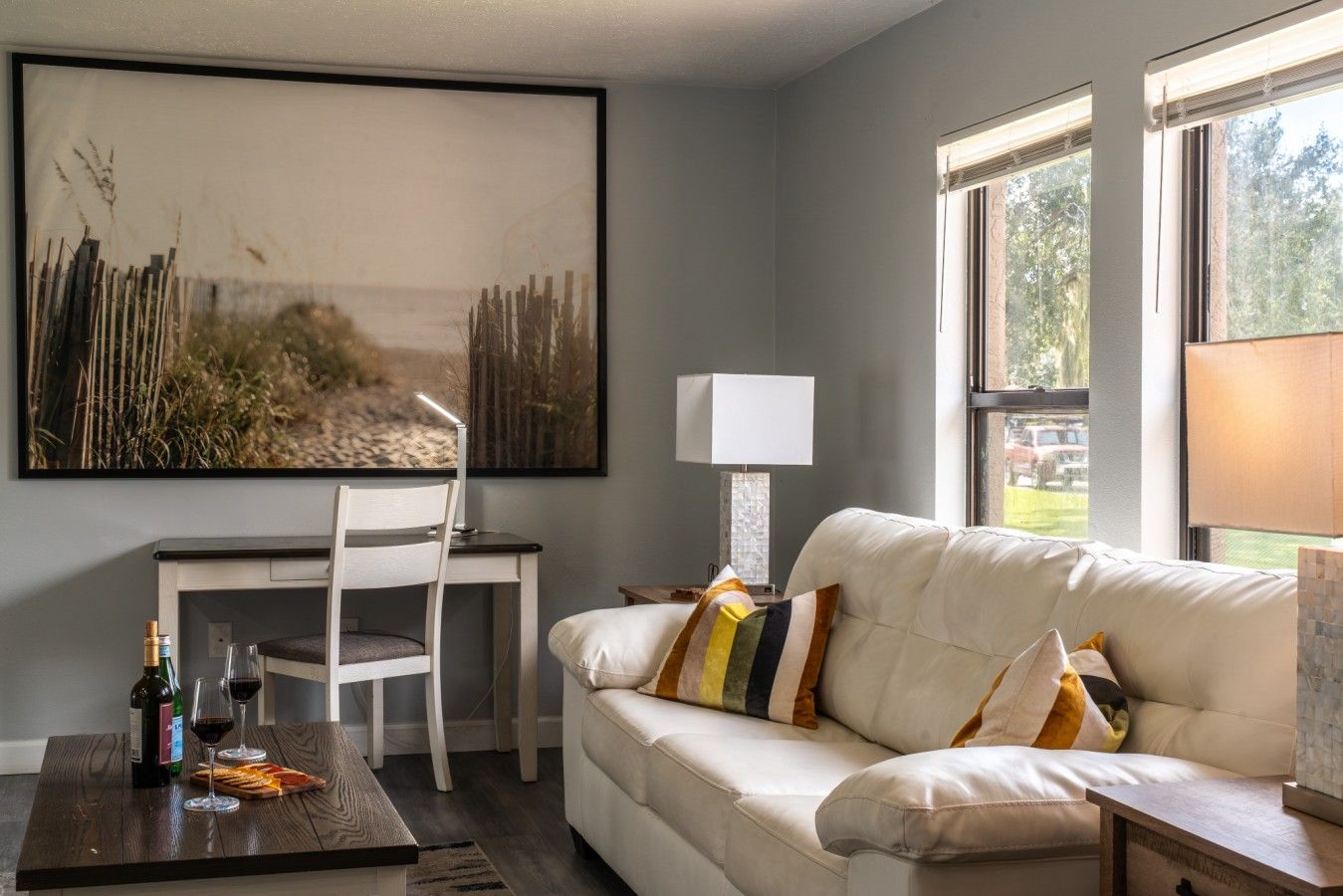 A living room with a white sofa, a dining table with chairs, a large painting, and a window with a view of trees.