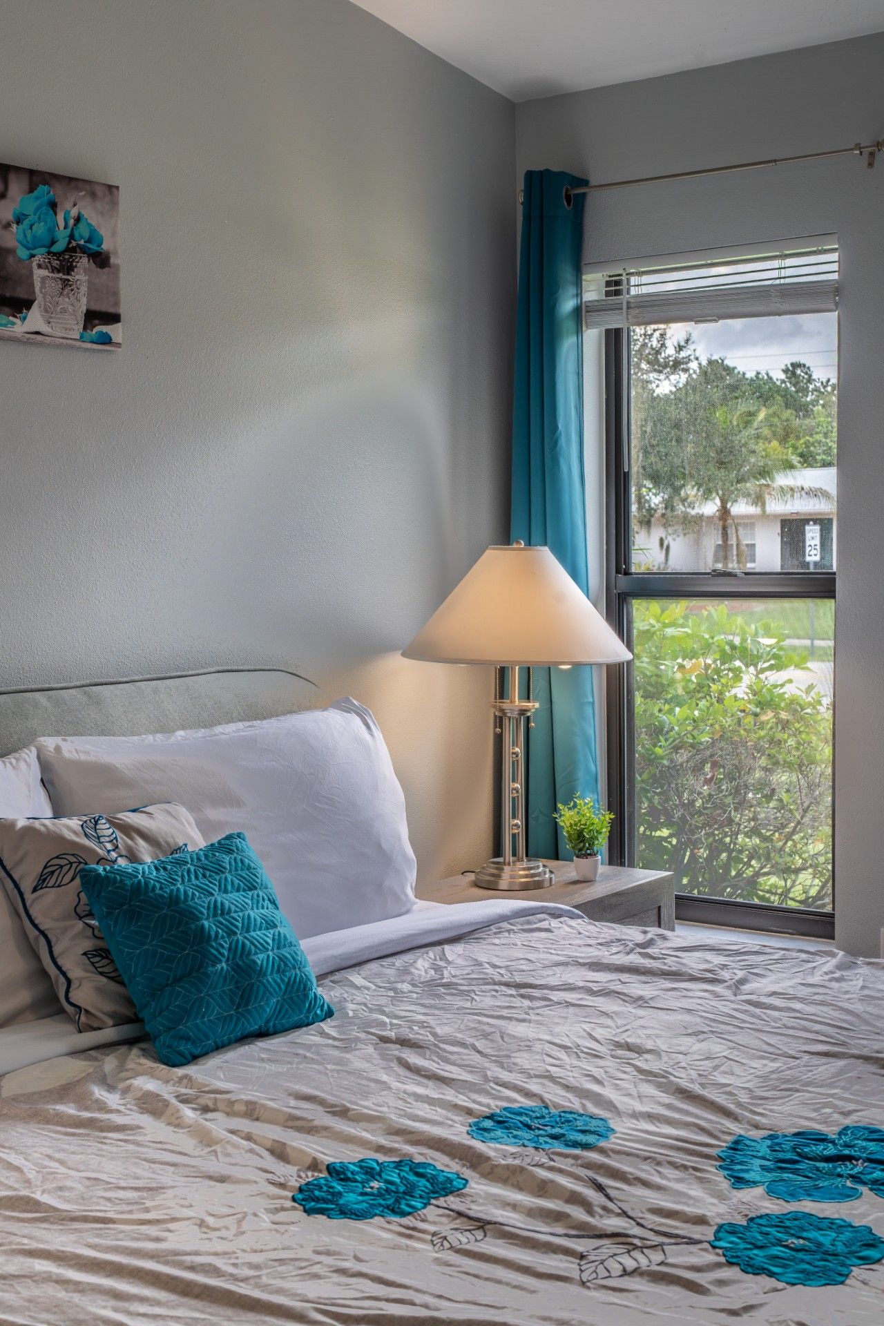 A bedroom with a bed, a bedside lamp, a window with curtains, and a view of greenery outside.