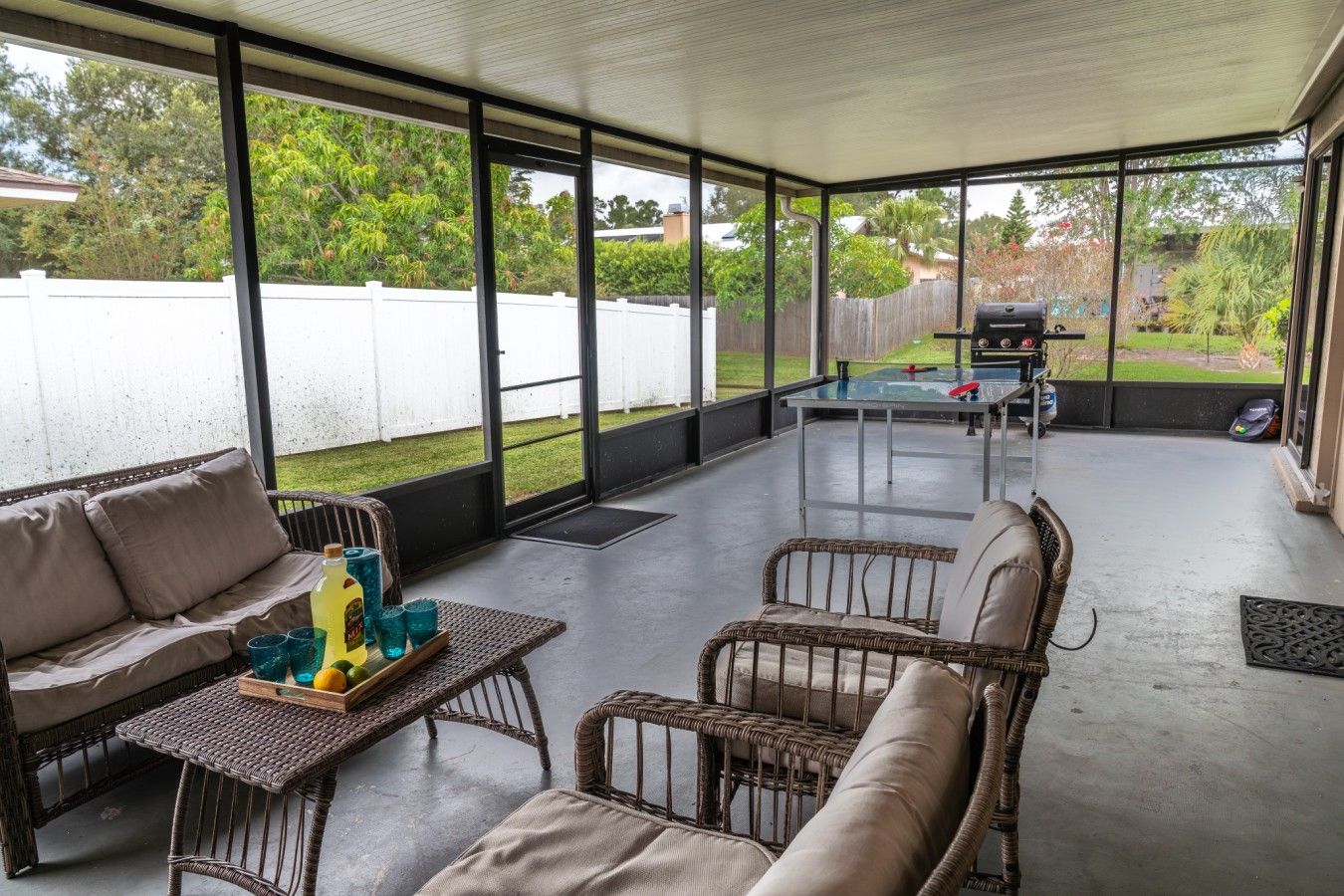 A covered patio with wicker furniture, a glass table, and a barbecue grill in the background.