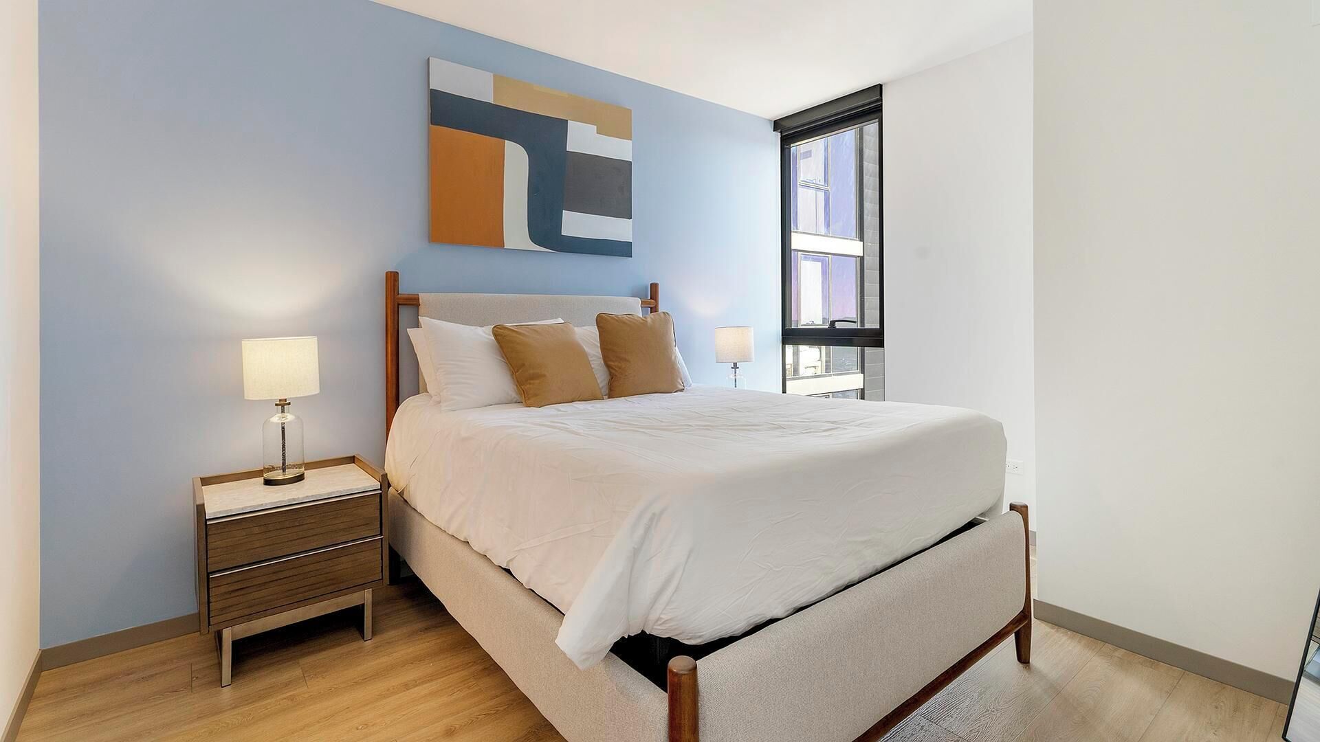 Un dormitorio con cama, mesitas de noche, lámpara, ventana y una obra de arte en la pared.