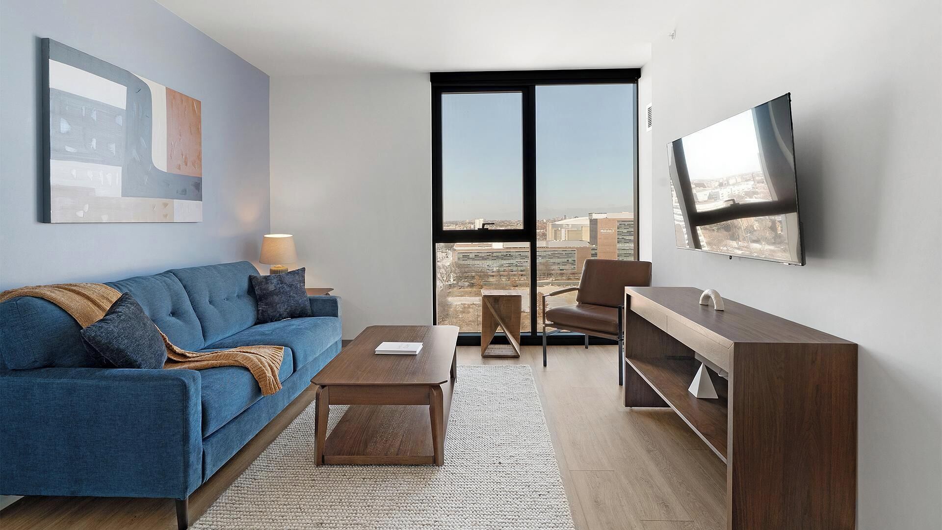 Un salon moderne avec un canapé, une table basse, un téléviseur et une vue sur la ville grâce à de grandes fenêtres.