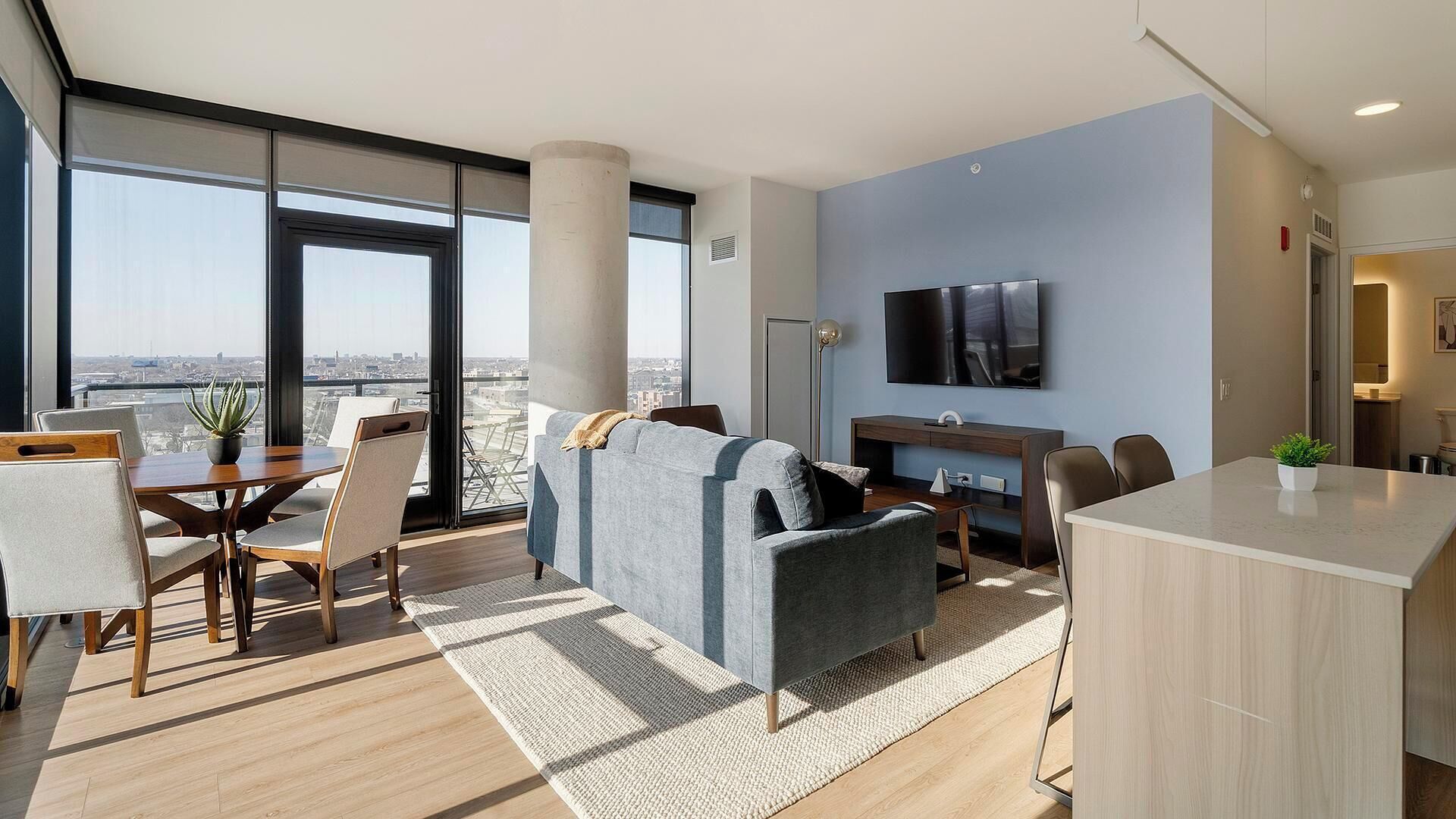 A modern hotel room with a large window, a flat-screen TV, a sofa, a dining table, and a kitchen area.