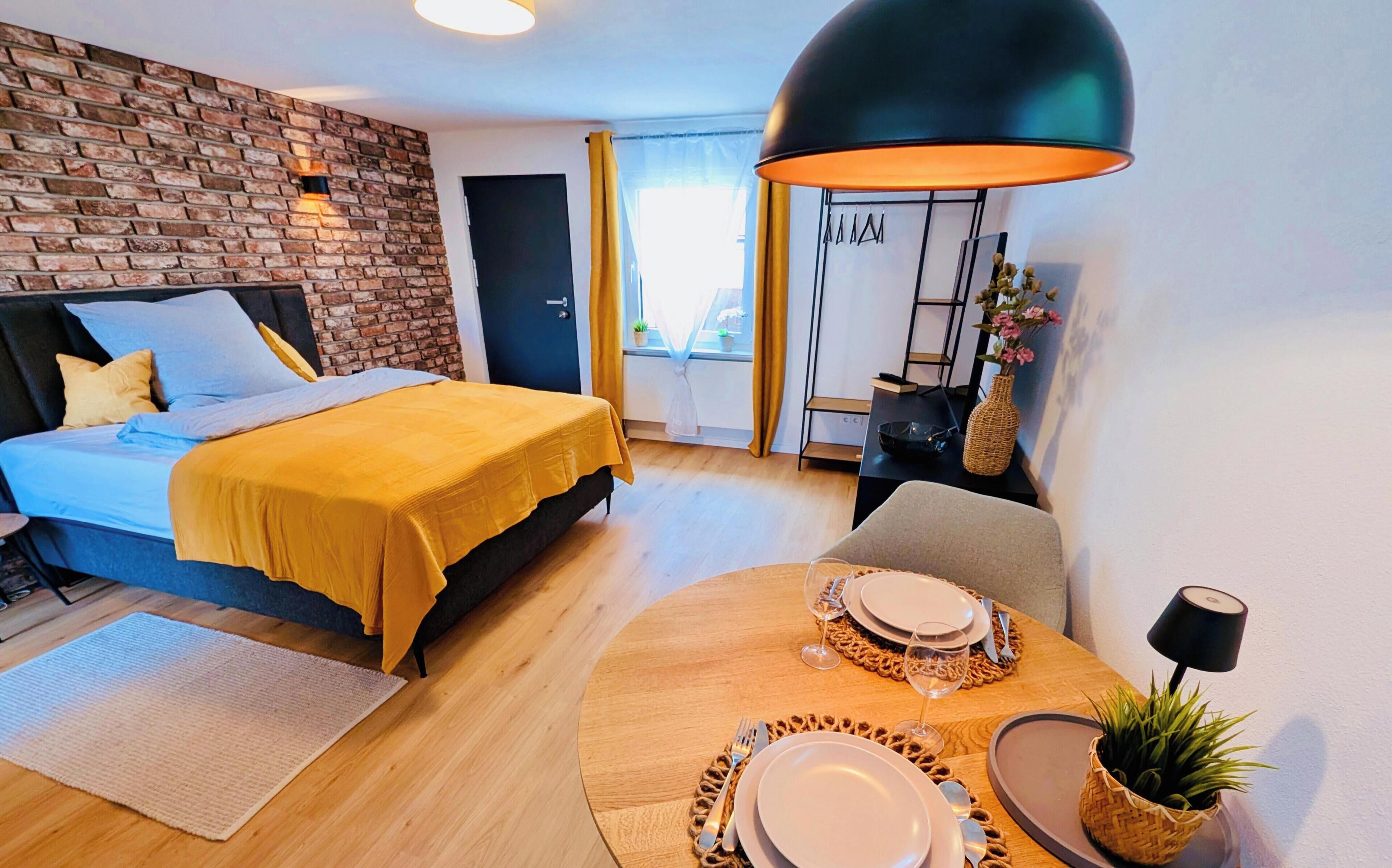 Una habitación de hotel moderna con una pared de ladrillo decorativa, una cama con ropa de cama amarilla, un rincón de comedor con una mesa redonda para dos personas y una gran lámpara colgante negra.