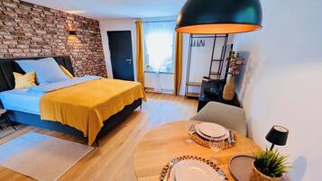 Una habitación de hotel moderna con una pared de ladrillo decorativa, una cama con ropa de cama amarilla, un rincón de comedor con una mesa redonda para dos personas y una gran lámpara colgante negra.