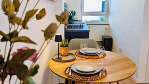 Uma sala de jantar com uma mesa de madeira redonda, cadeiras pretas, uma luminária pendente preta e vista para a cozinha ao fundo.