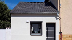 Um pequeno edifício térreo com telhado de telhas, uma janela com cortina e uma porta com puxador.