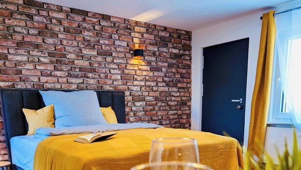 Quarto de hotel com parede de tijolos, cama com roupa de cama amarela, mesa de cabeceira com um copo de vinho e uma planta em vaso.
