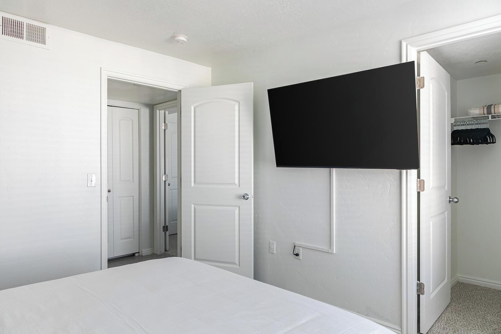Ein Schlafzimmer mit einem Bett, einem an der Wand montierten Fernseher und einem Kleiderschrank mit Kleidung.