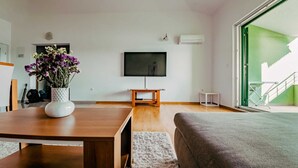 A living room with a wooden table, a TV on the wall, a small white side table, and a large window with a view of a building. - Seaview apartment Verde near beach (Okrug Gornji)