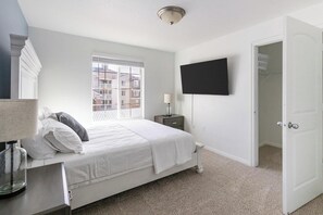 Ein Schlafzimmer mit einem Bett, einem Nachttisch mit Lampe, einem an der Wand befestigten Fernseher und einem Fenster mit Blick auf Gebäude.