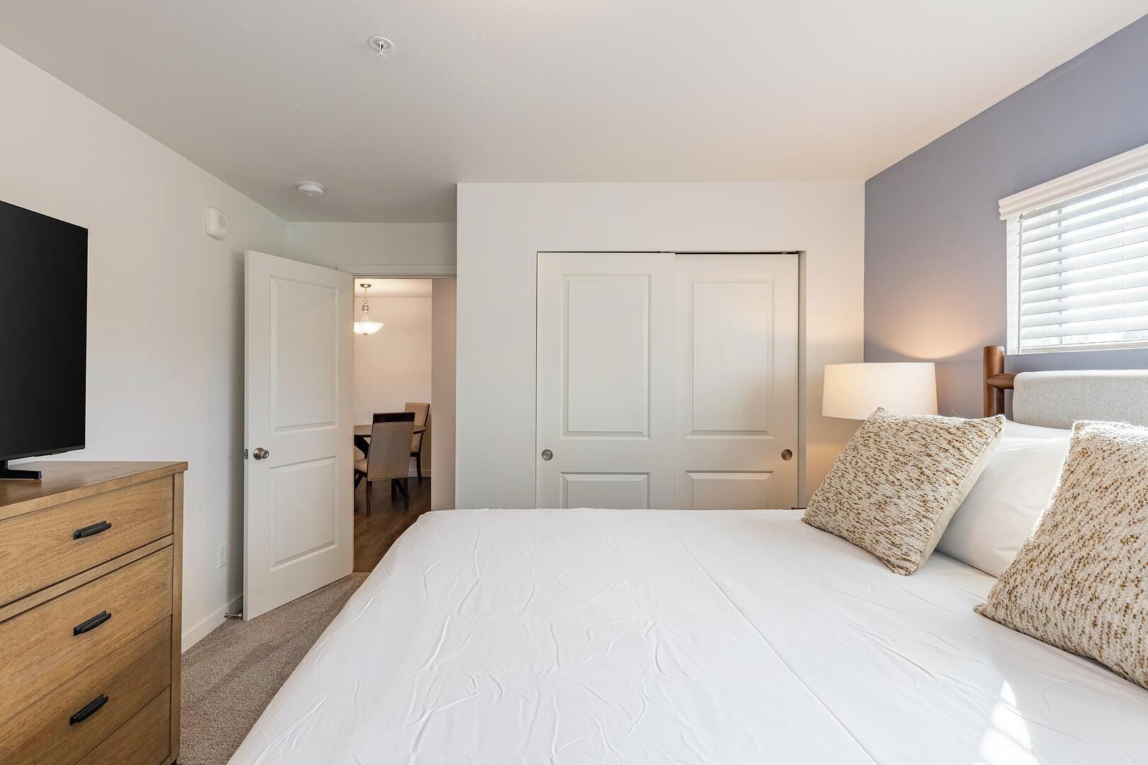 A bedroom with a bed, a dresser, a television, a door, and a window with blinds.