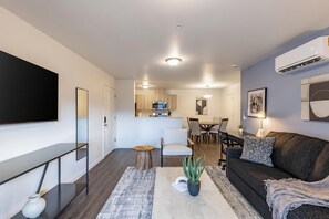 A modern living room with a flat-screen TV, a dark-colored sofa, a coffee table, and a dining area with chairs and a table. - Landing | Amazing 2BD, Clubhouse, Gym (Richland)