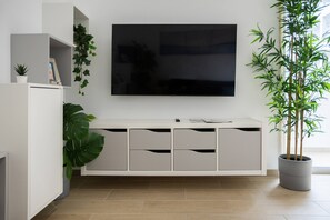 A modern living room with a flat-screen TV mounted on the wall, a white entertainment unit with drawers, a large green plant, and a potted bamboo plant. - Pé na Areia by Centralgarve (Loulé)