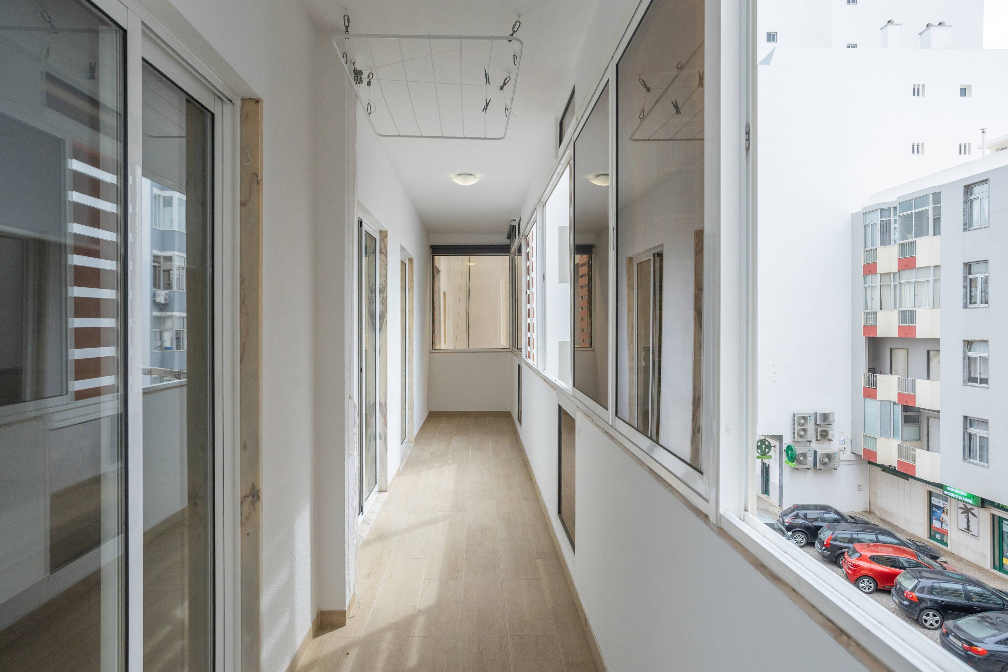 A narrow corridor with a wooden floor, large windows, and a view of parked cars outside.