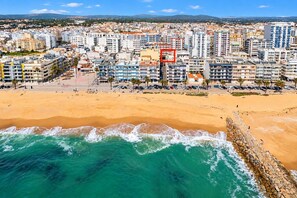 A coastal cityscape with a sandy beach, a rocky breakwater, and a dense urban area with multi-story buildings. - "Pé na Areia" by Centralgarve (Loulé)