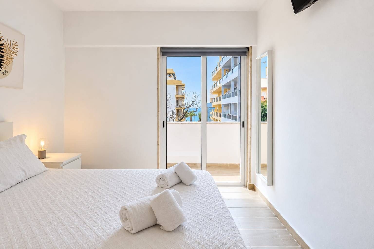 Une chambre d’hôtel avec un lit, une table de chevet, une lampe et une vue sur un immeuble, visible à travers une porte coulissante.