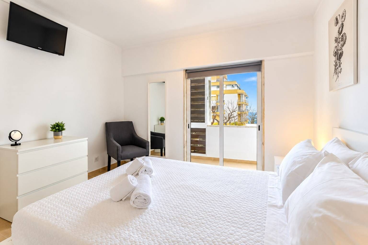 A hotel room with a bed, a chair, a desk with a plant, a flat-screen TV, and a balcony with a view.