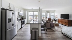 Eine moderne Küche mit Edelstahlgeräten, ein Essbereich mit einer zentralen Kochinsel und ein Wohnbereich mit Sofa und Fernseher.
