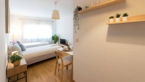 Ein modernes Wohnzimmer mit einer Couch, einem Esstisch und einem Computer. Regale mit Pflanzen und eine hängende Pflanze sind ebenfalls vorhanden.